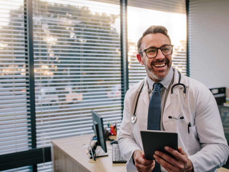 Smiling mature male doctor with digital tablet in his office. Friendly medical professional with tablet computer in clinic.